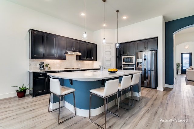 kitchen featuring backsplash, appliances with stainless steel finishes, decorative light fixtures, an island with sink, and light hardwood / wood-style floors