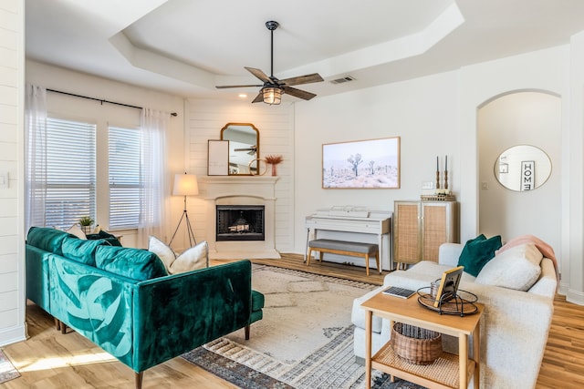 living room with a large fireplace, ceiling fan, a raised ceiling, and light hardwood / wood-style flooring