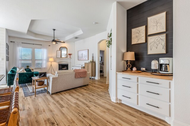 living room with ceiling fan, a large fireplace, light hardwood / wood-style floors, and a raised ceiling