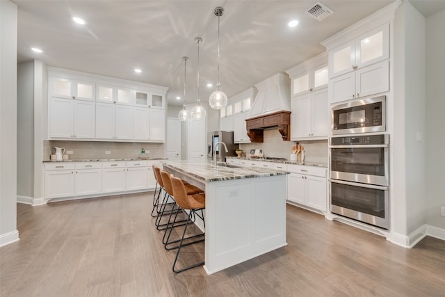 kitchen featuring white cabinets, custom range hood, an island with sink, and stainless steel appliances