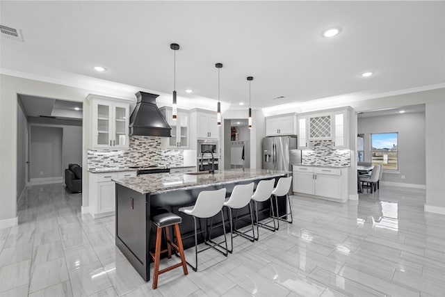 kitchen with stainless steel appliances, white cabinetry, custom exhaust hood, decorative light fixtures, and a kitchen island with sink