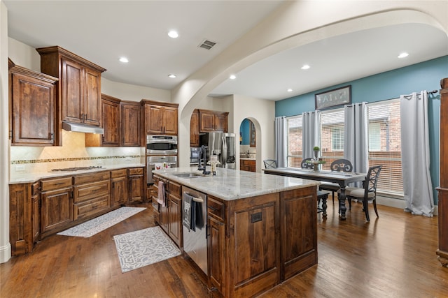 kitchen with stainless steel appliances, dark hardwood / wood-style floors, light stone countertops, sink, and an island with sink