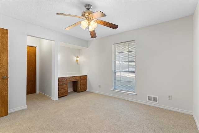 unfurnished bedroom with built in desk, ceiling fan, a textured ceiling, and light colored carpet