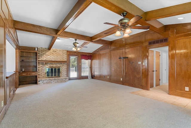 unfurnished living room with wood walls, light carpet, ceiling fan, beam ceiling, and a brick fireplace