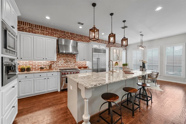 kitchen with an island with sink, built in appliances, white cabinets, and wall chimney range hood