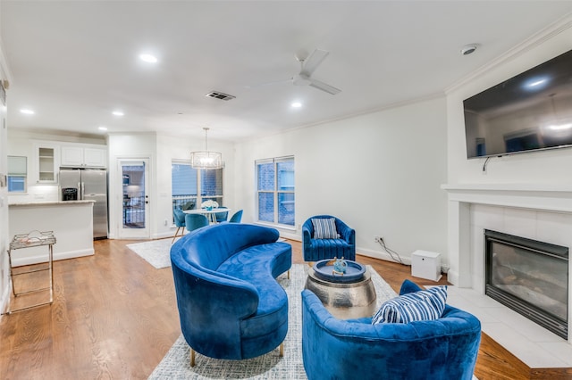 living room with a tiled fireplace, light hardwood / wood-style flooring, ceiling fan with notable chandelier, and ornamental molding