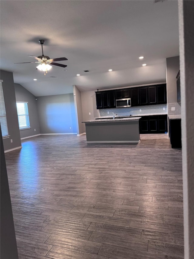 kitchen with dark wood-type flooring, a center island with sink, backsplash, and ceiling fan