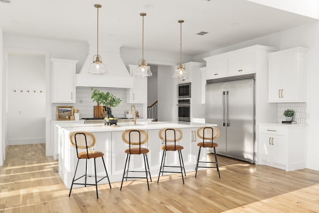 kitchen featuring built in appliances, a kitchen island with sink, white cabinetry, and backsplash