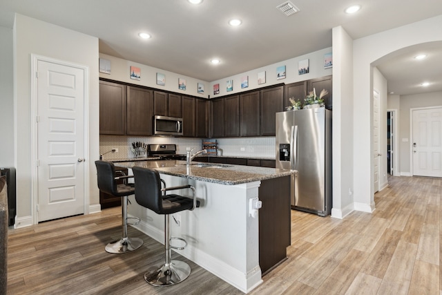 kitchen featuring light wood-type flooring, appliances with stainless steel finishes, a kitchen bar, dark brown cabinets, and an island with sink