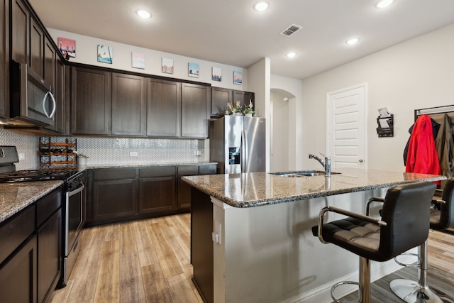 kitchen featuring stainless steel appliances, dark stone counters, sink, an island with sink, and light wood-type flooring