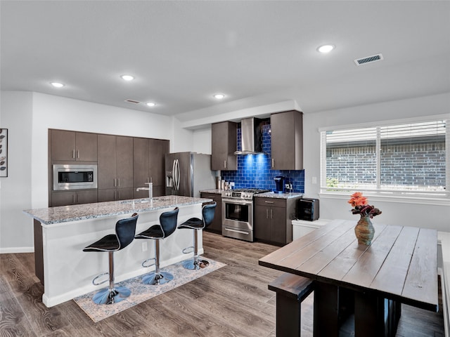 kitchen featuring appliances with stainless steel finishes, a center island with sink, wall chimney range hood, and light hardwood / wood-style flooring