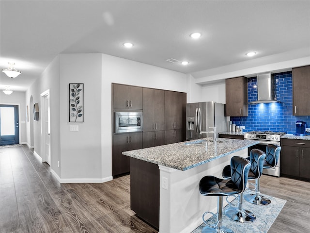 kitchen with appliances with stainless steel finishes, a kitchen breakfast bar, an island with sink, light hardwood / wood-style flooring, and wall chimney range hood