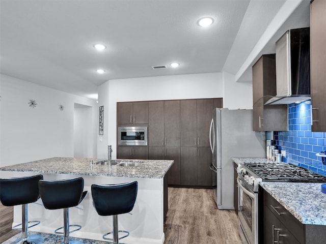 kitchen with wall chimney range hood, appliances with stainless steel finishes, sink, light hardwood / wood-style floors, and a breakfast bar