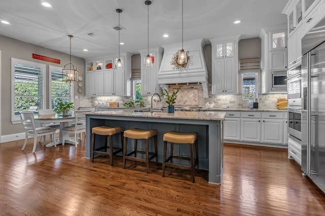 kitchen with dark wood-type flooring, a center island with sink, decorative backsplash, light stone countertops, and white cabinetry