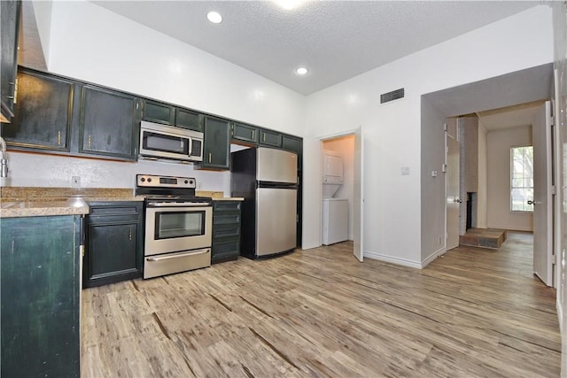 kitchen with appliances with stainless steel finishes, light stone countertops, a textured ceiling, light hardwood / wood-style flooring, and stacked washer and dryer