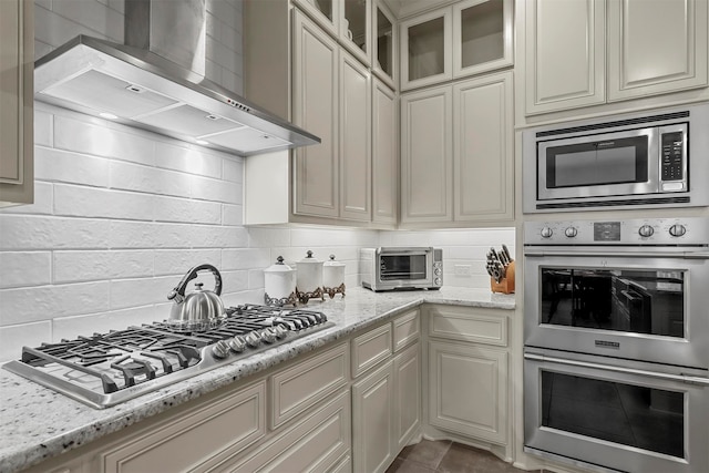 kitchen with tasteful backsplash, stainless steel appliances, wall chimney range hood, light stone countertops, and cream cabinetry