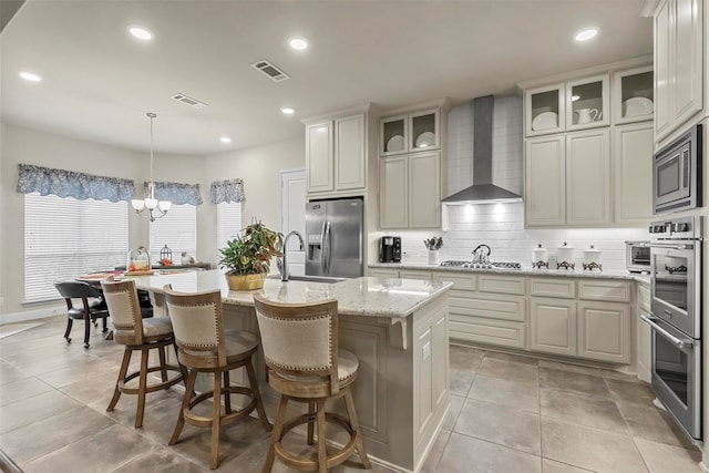 kitchen with appliances with stainless steel finishes, an inviting chandelier, decorative light fixtures, an island with sink, and wall chimney range hood