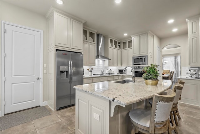 kitchen featuring stainless steel appliances, a center island with sink, a kitchen breakfast bar, wall chimney range hood, and light stone countertops