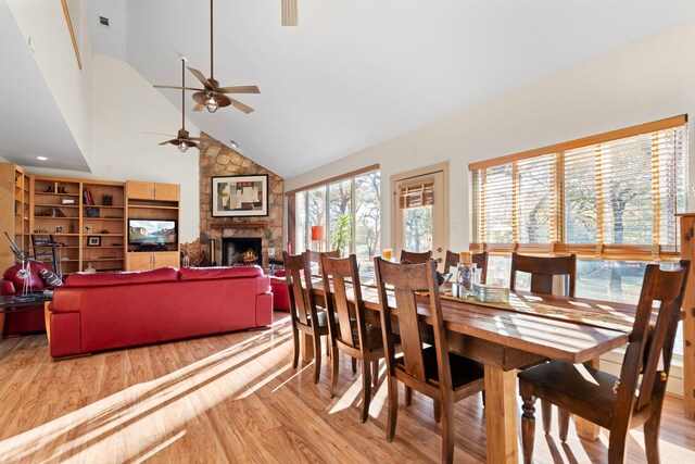 dining area with a stone fireplace, ceiling fan, high vaulted ceiling, and light wood-type flooring