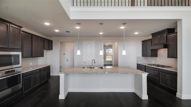 kitchen featuring stainless steel appliances, a center island with sink, sink, light stone countertops, and dark wood-type flooring