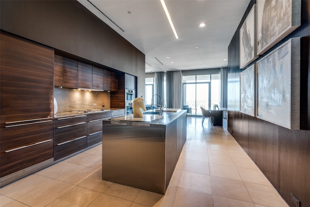 kitchen featuring dark brown cabinetry, sink, an island with sink, light tile patterned flooring, and backsplash