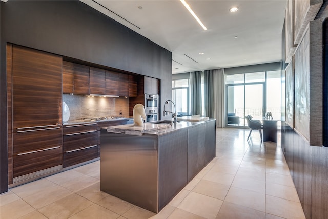 kitchen featuring light stone counters, light tile patterned floors, decorative backsplash, sink, and an island with sink