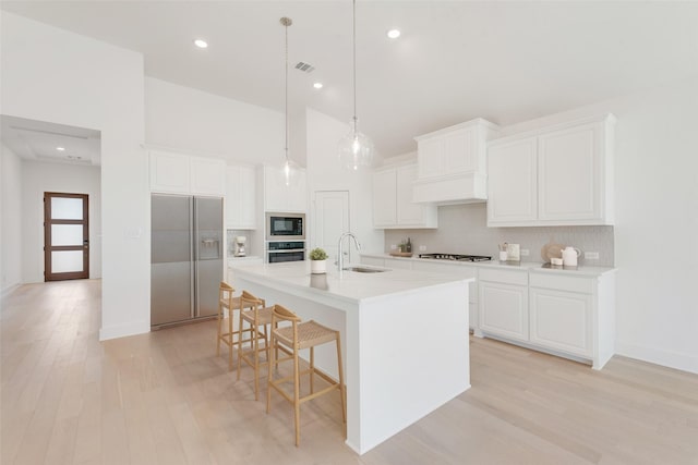 kitchen with sink, white cabinetry, built in appliances, hanging light fixtures, and a kitchen island with sink