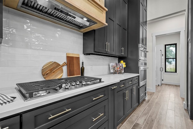 kitchen with appliances with stainless steel finishes, light hardwood / wood-style flooring, decorative backsplash, and exhaust hood