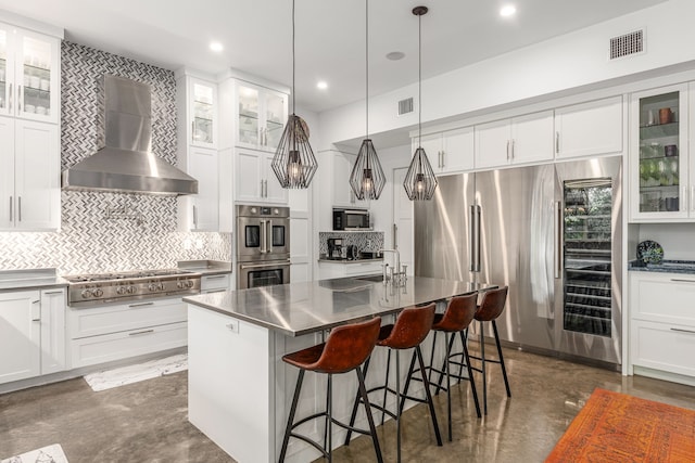 kitchen with hanging light fixtures, a breakfast bar area, a kitchen island with sink, wall chimney range hood, and appliances with stainless steel finishes