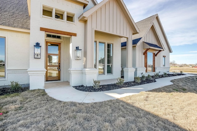 doorway to property featuring board and batten siding, a yard, brick siding, and a shingled roof