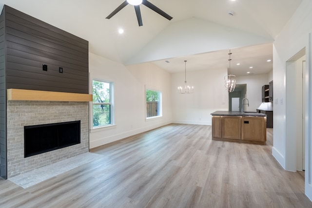 unfurnished living room with ceiling fan with notable chandelier, sink, high vaulted ceiling, a fireplace, and light hardwood / wood-style flooring