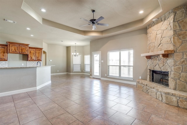 unfurnished living room featuring a stone fireplace, ceiling fan with notable chandelier, sink, light tile patterned floors, and a raised ceiling