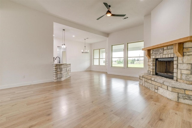 unfurnished living room with a stone fireplace, sink, ceiling fan with notable chandelier, and light wood-type flooring