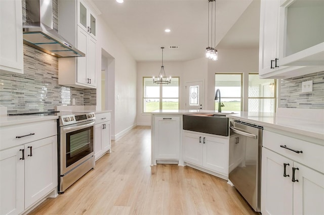 kitchen with sink, hanging light fixtures, stainless steel appliances, wall chimney range hood, and white cabinets