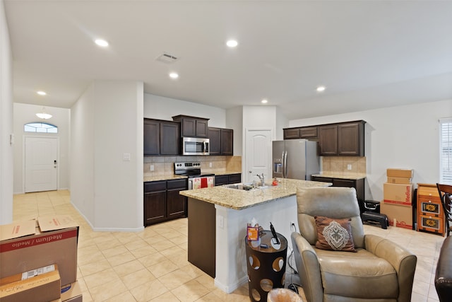 kitchen with appliances with stainless steel finishes, dark brown cabinets, sink, decorative backsplash, and an island with sink