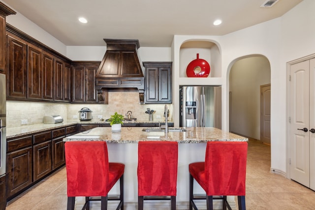 kitchen featuring custom exhaust hood, stainless steel fridge, a kitchen island with sink, light stone counters, and sink