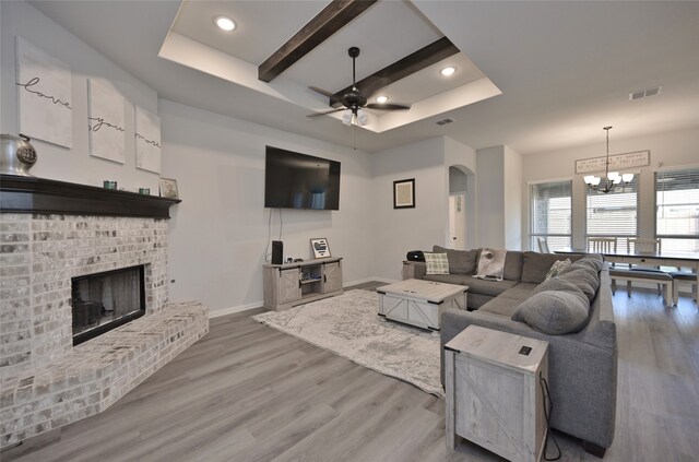 living room featuring wood-type flooring, beam ceiling, a brick fireplace, a tray ceiling, and ceiling fan with notable chandelier