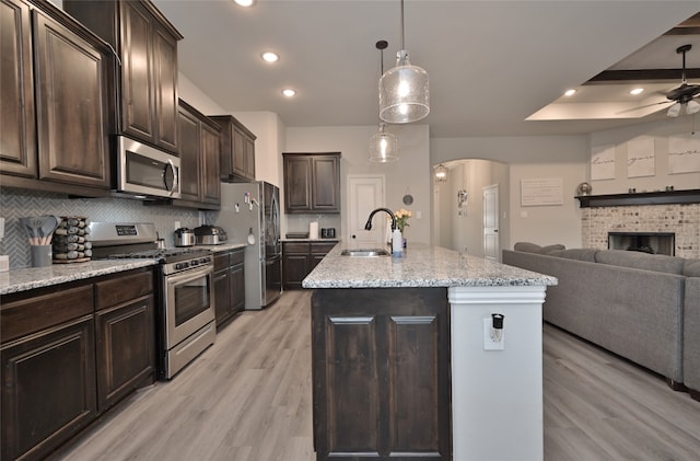 kitchen featuring a brick fireplace, light hardwood / wood-style flooring, an island with sink, appliances with stainless steel finishes, and decorative light fixtures