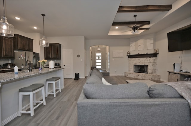 living room with a brick fireplace, light hardwood / wood-style floors, sink, beamed ceiling, and ceiling fan