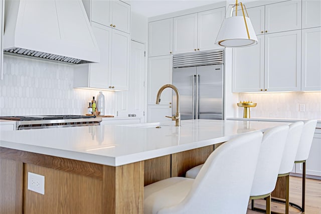 kitchen with built in fridge, custom exhaust hood, light countertops, decorative backsplash, and a sink