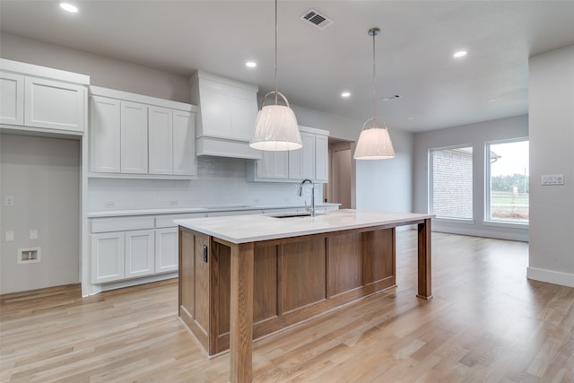 kitchen with pendant lighting, a kitchen island with sink, white cabinets, sink, and light hardwood / wood-style flooring