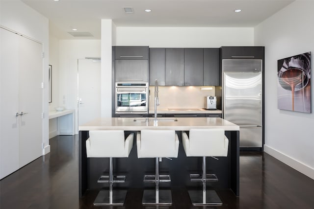 kitchen featuring sink, appliances with stainless steel finishes, a kitchen bar, a kitchen island with sink, and dark wood-type flooring