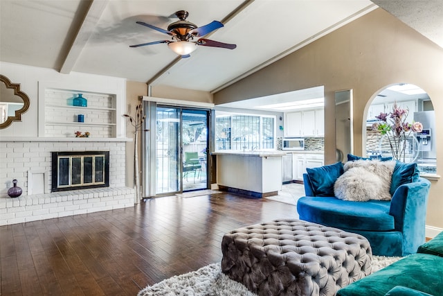 living room featuring ceiling fan, wood-type flooring, vaulted ceiling with beams, and a brick fireplace