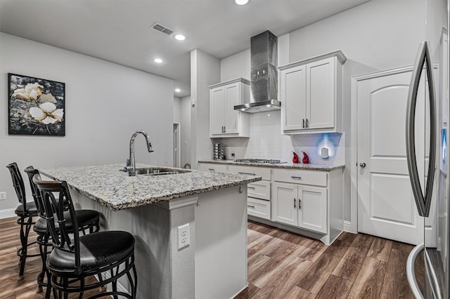 kitchen with a kitchen island with sink, white cabinets, a kitchen breakfast bar, sink, and wall chimney exhaust hood