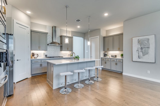 kitchen featuring gray cabinets, decorative light fixtures, an island with sink, a breakfast bar area, and wall chimney range hood