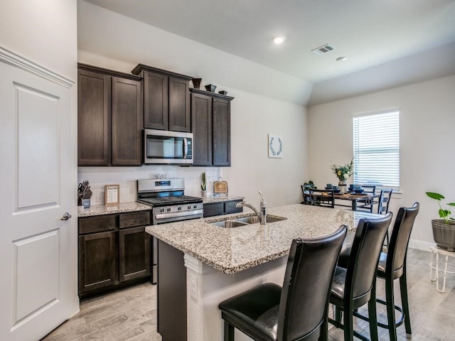 kitchen with stainless steel appliances, sink, light stone counters, a kitchen island with sink, and light hardwood / wood-style flooring