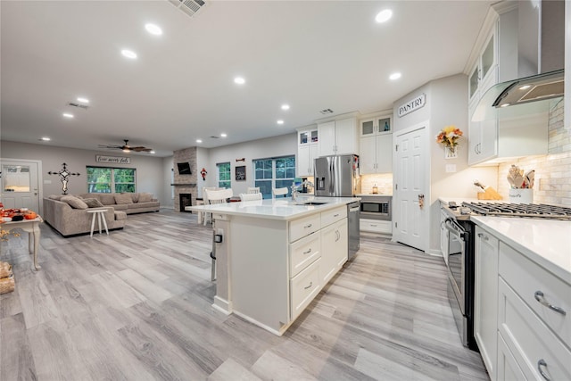 kitchen featuring a fireplace, white cabinets, stainless steel appliances, a center island with sink, and wall chimney exhaust hood