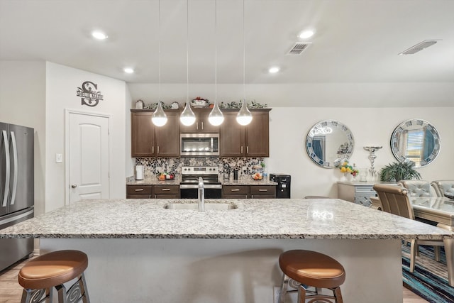 kitchen with appliances with stainless steel finishes, light hardwood / wood-style floors, decorative light fixtures, and an island with sink