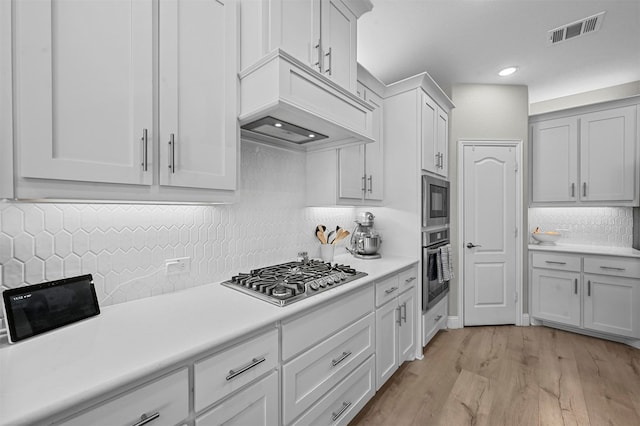 kitchen with premium range hood, light wood-type flooring, appliances with stainless steel finishes, decorative backsplash, and white cabinets