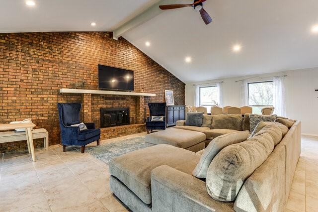 living room with ceiling fan, brick wall, a brick fireplace, and lofted ceiling with beams